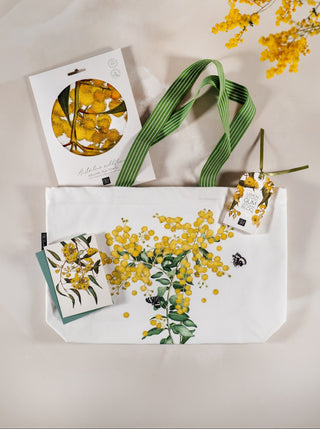 A white canvas tote bag with a yellow and green glowing wattle floral print, with a green striped handle, a lemon scented gum floral card, a golden wattle organic linen and cotton tea towel and a clothes protecting lemon scented gum aroma bloq on a light beige background.